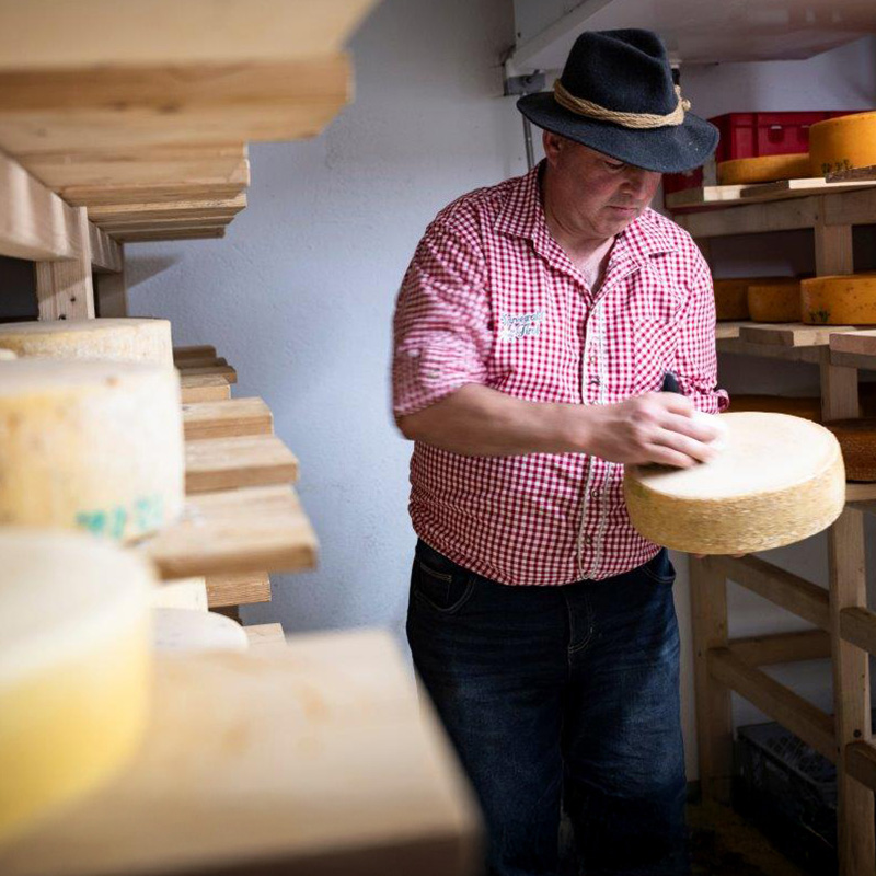 Marcel Schallmea prüft ein Käselaib im Reifekeller der Hofkäserei, im Hintergrund Käseleiber auf Holzregalen.